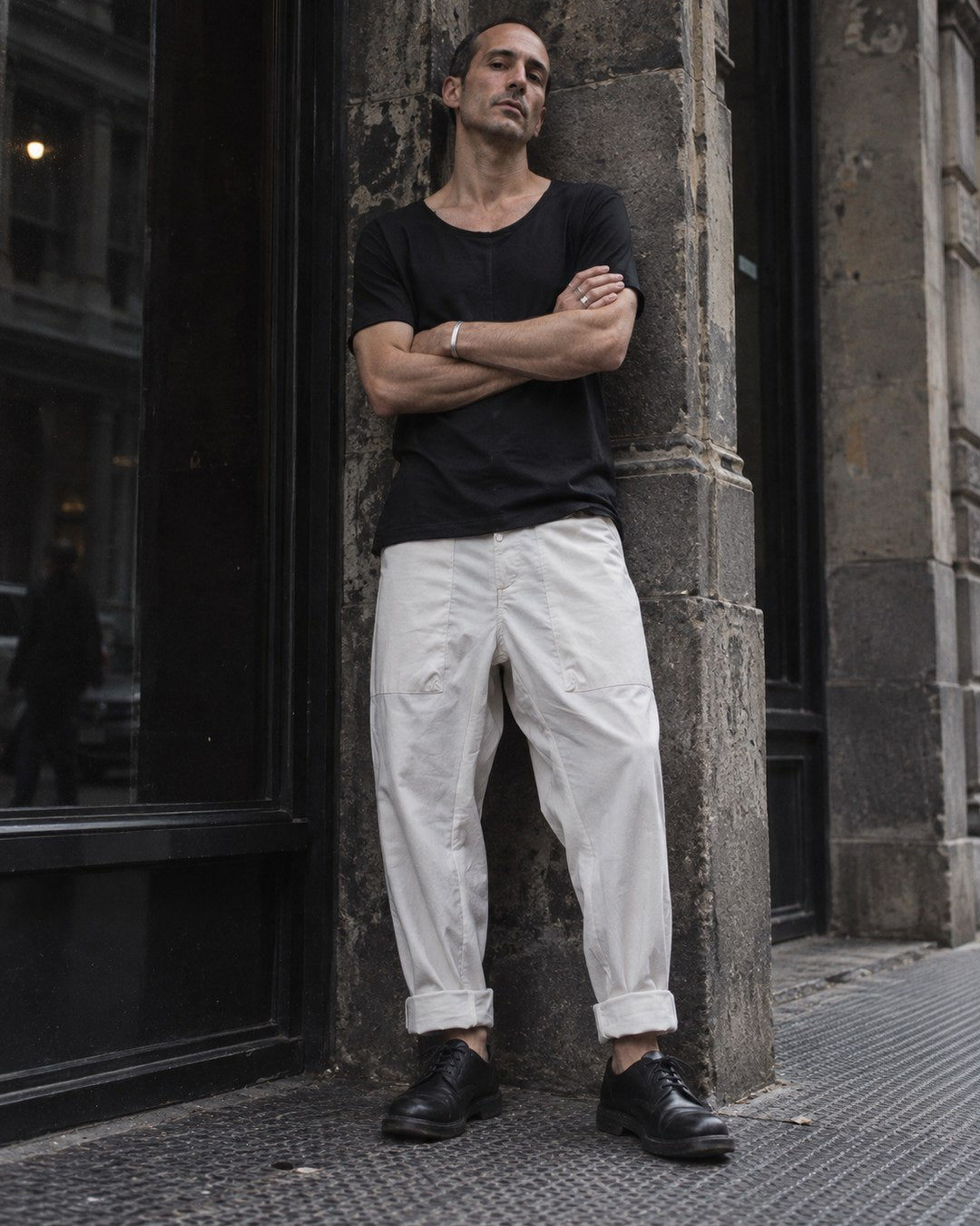 AITO scoop neck cotton t-shirt in black with relaxed cotton trousers in off white, full length, in a doorway on the street