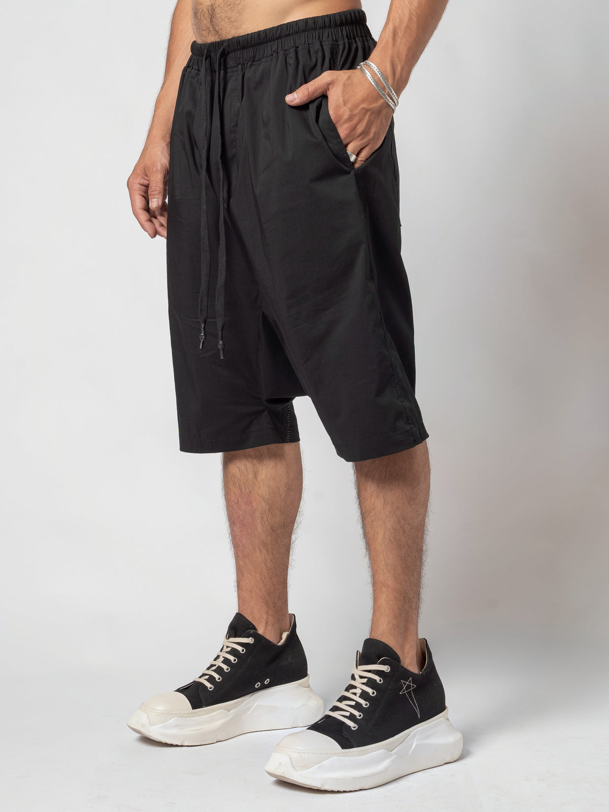 AITO Drop-Crotch Cotton Shorts in Black — three-quarter front view with one hand slipped into the side pocket and the drawcord hanging on the near hip