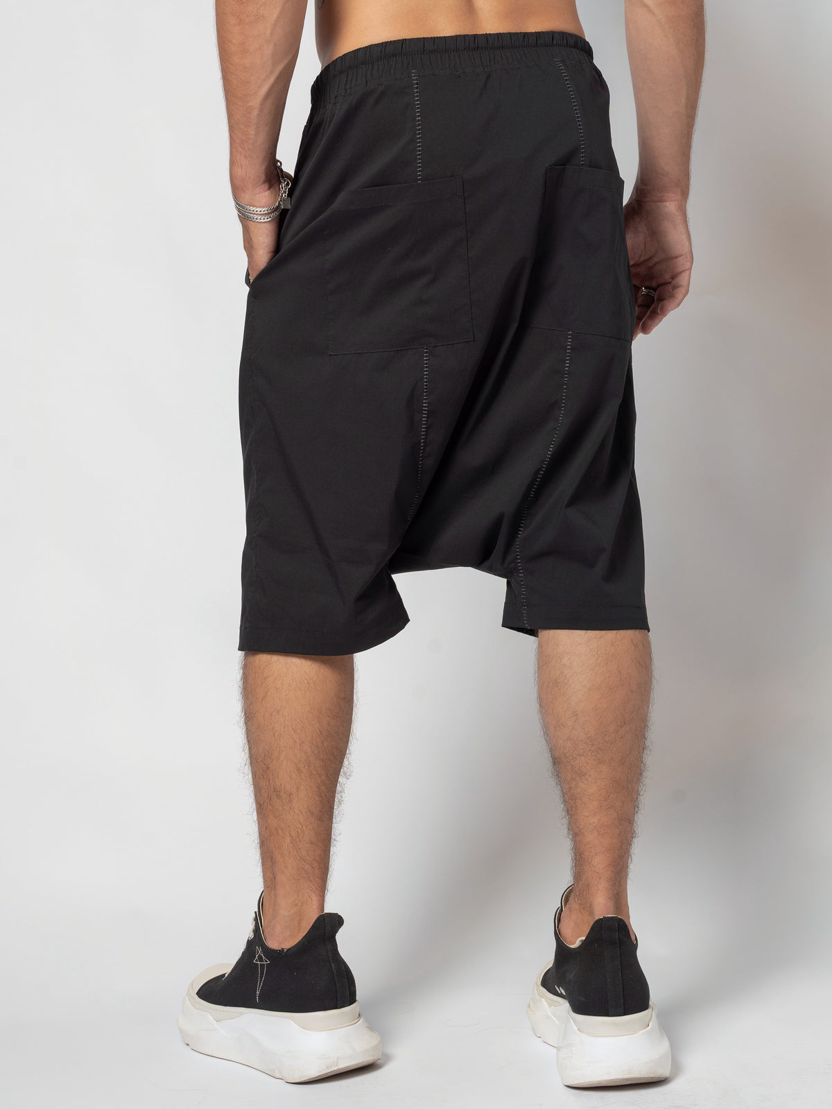 AITO Drop-Crotch Cotton Shorts in Black — back view with one hand tucked at the hip and the other arm hanging free, feet planted wide apart