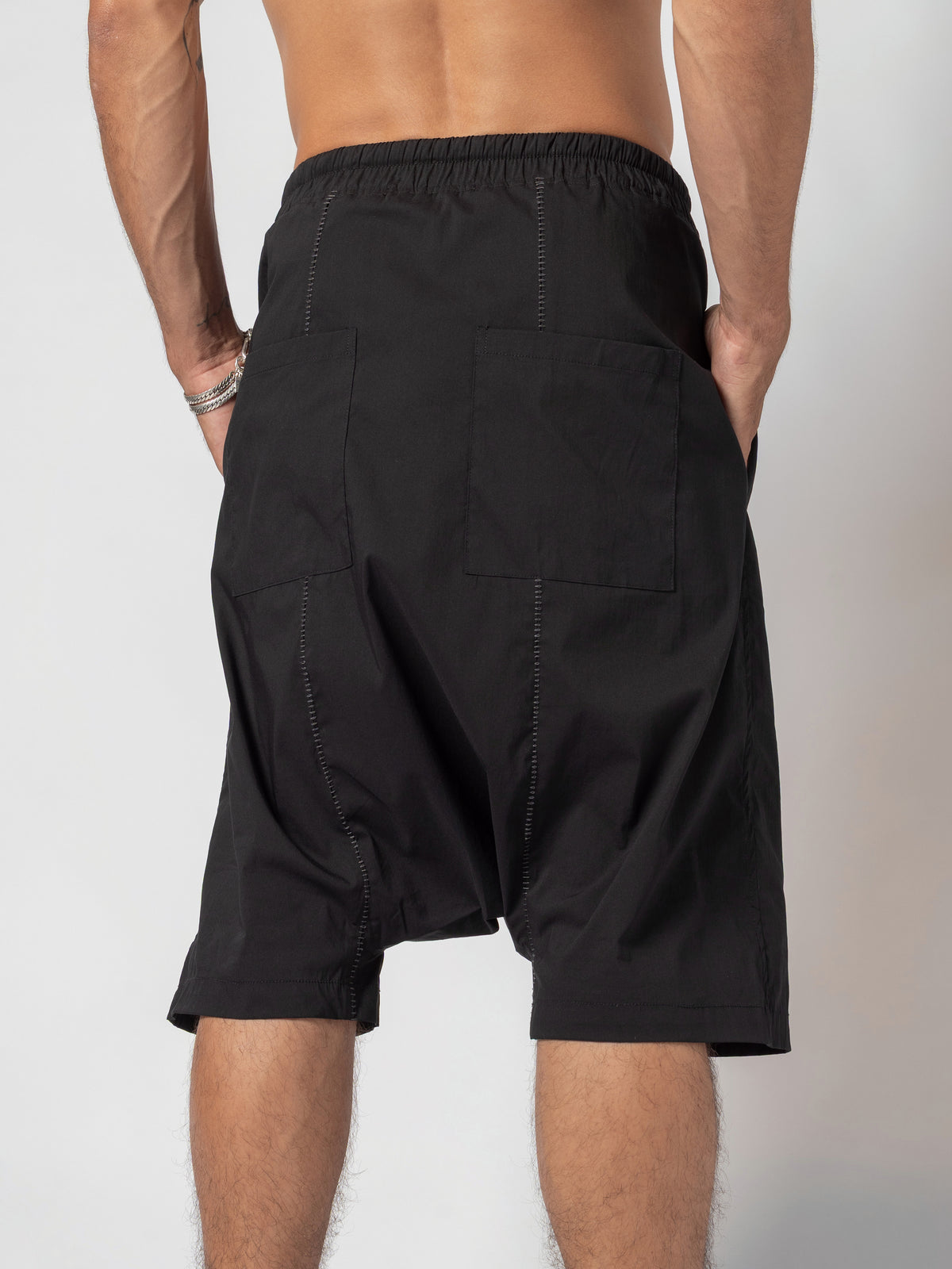 AITO Drop-Crotch Cotton Shorts in Black — back view cropped at the knee, both hands slipped into the pockets and the seat hanging low between the legs