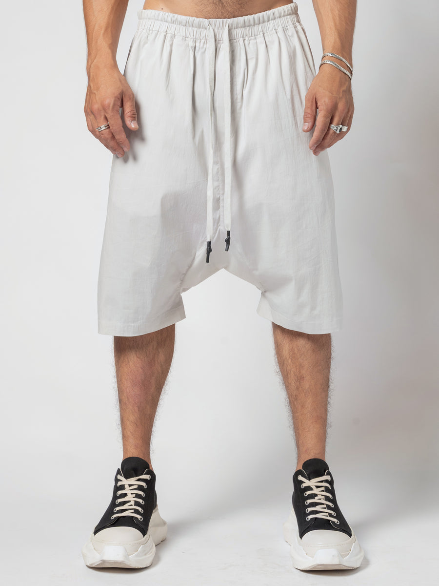 AITO Drop-Crotch Cotton Shorts in Off White — front view from the waist down, both hands at the sides, drawcord hanging over the low crotch, worn with black trainers