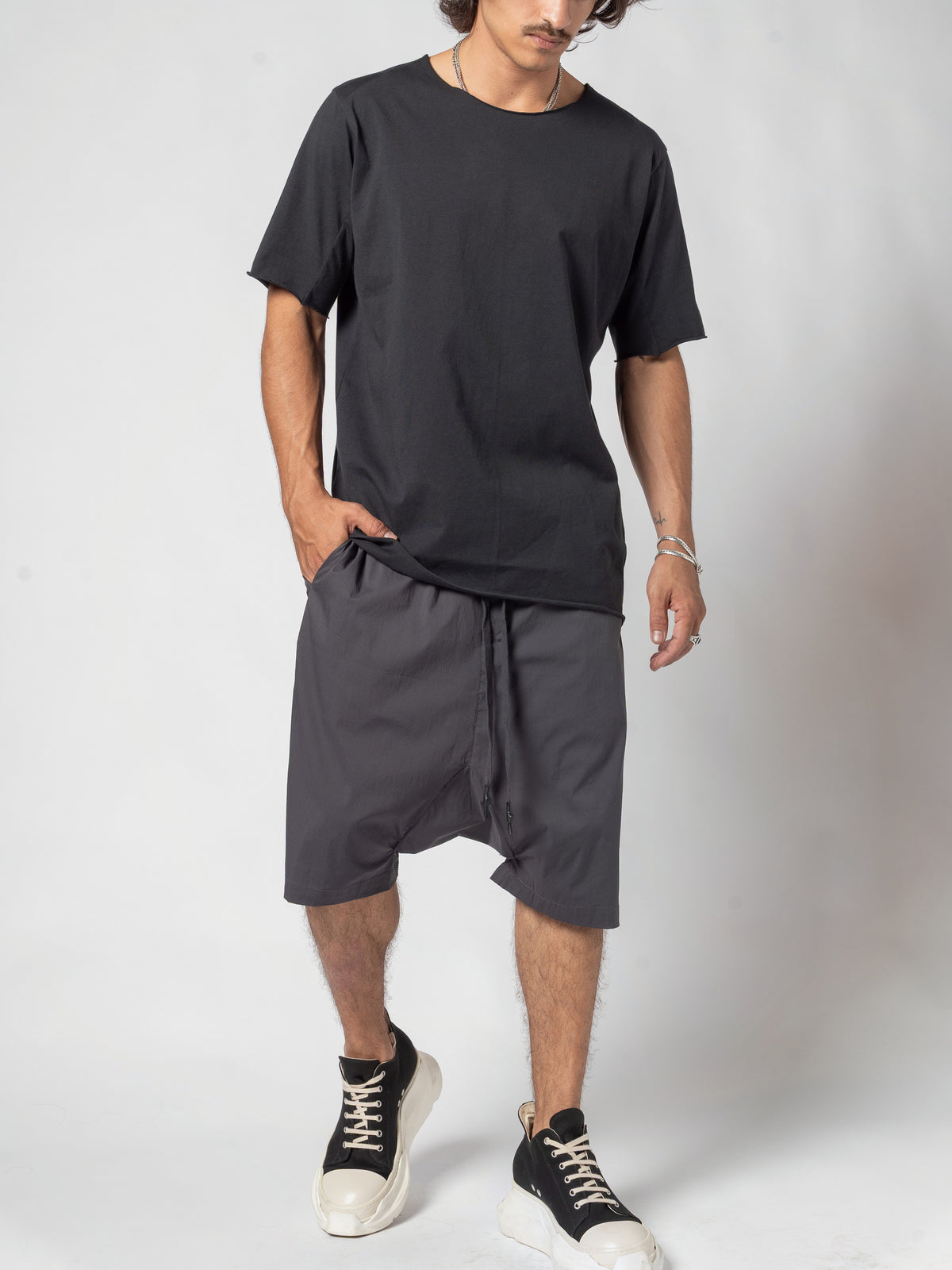 AITO Scar Stitch Cotton T-Shirt in Black — full-length front view with one hand in the shorts pocket, the hem lifted on that side