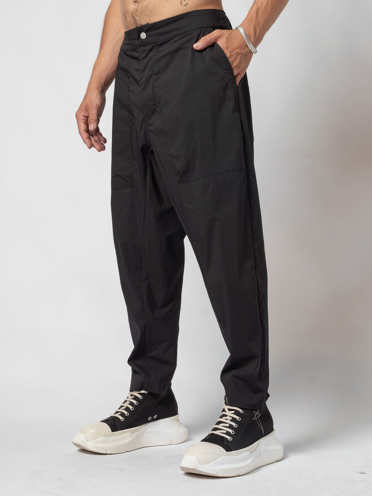 AITO Pleated Drop-Crotch Pants in Black — front three-quarter with one hand in the side pocket, feet staggered and the hem stacking over the trainer