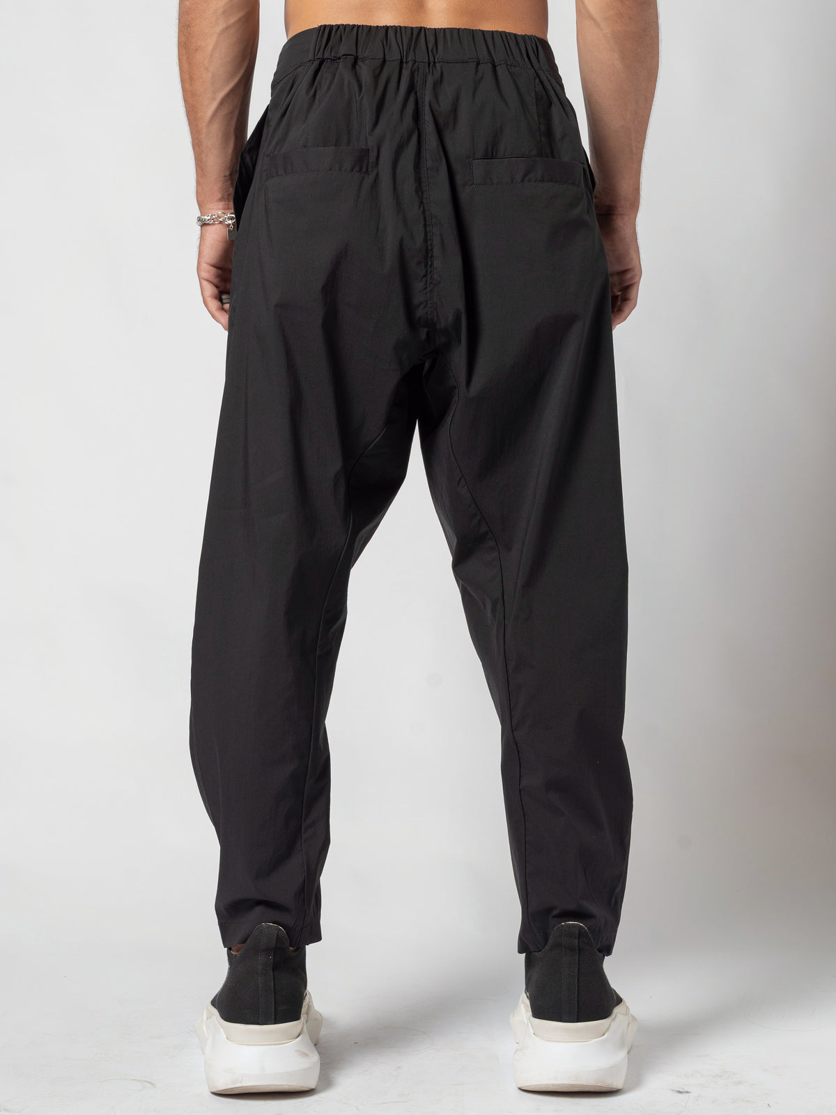 AITO Pleated Drop-Crotch Pants in Black — back view straight on, cropped to the gathered elastic waistband and the tapered legs, feet apart