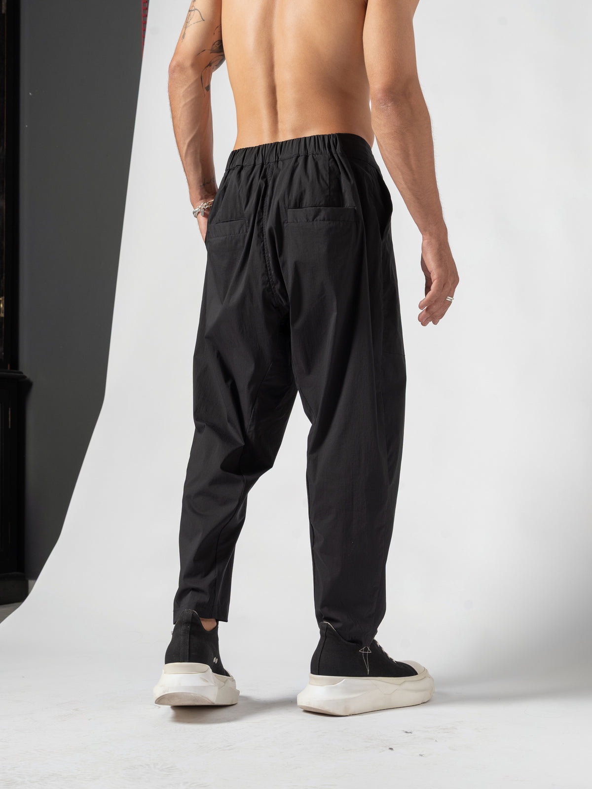 AITO Pleated Drop-Crotch Pants in Black — back three-quarter from further away, the free arm swung clear of the body and the far leg set back